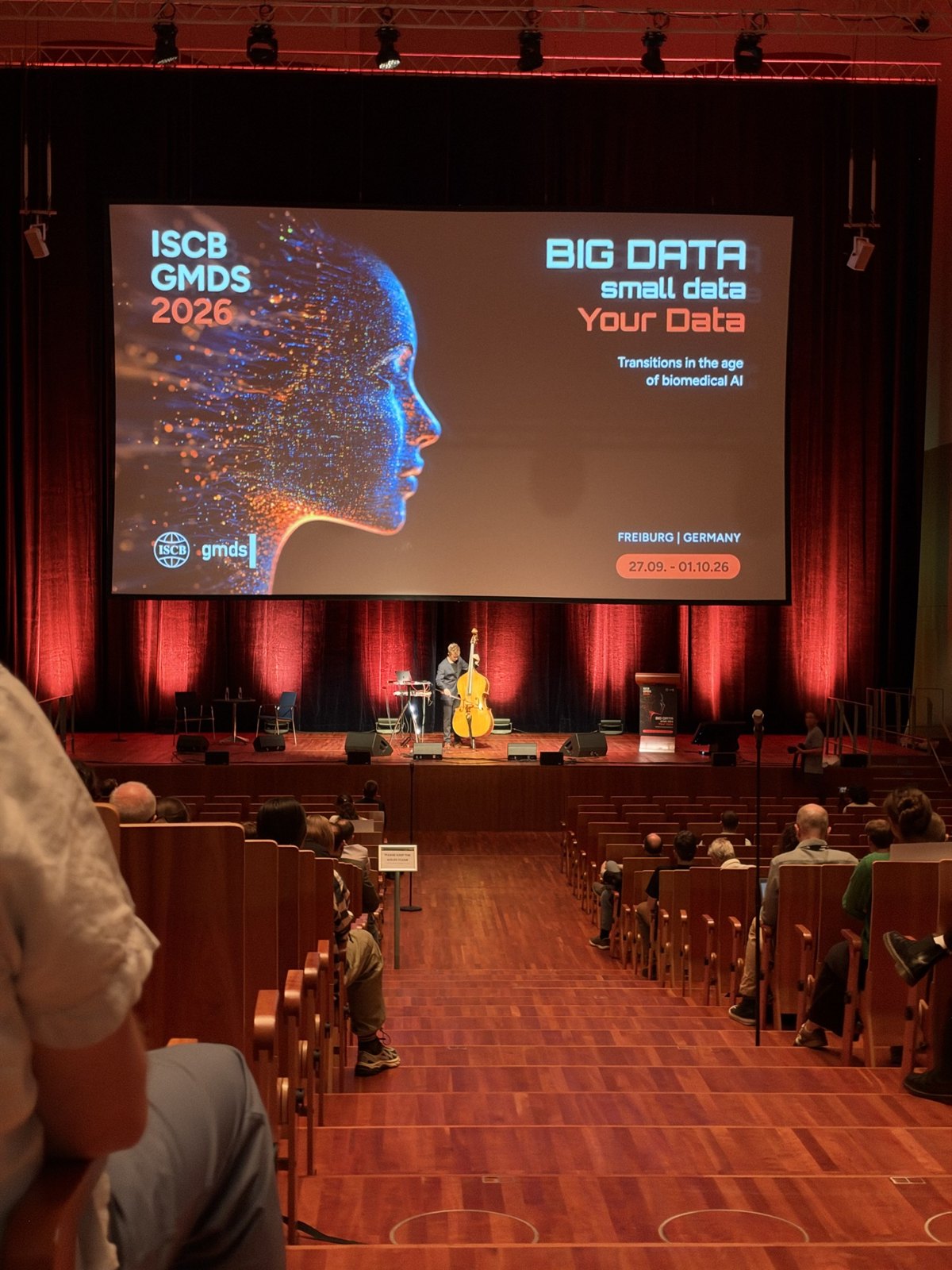 A double bassist on the Konzerthaus stage beneath the ISCB GMDS 2026 conference screen.